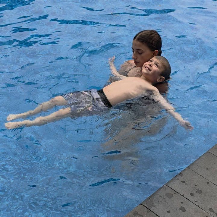 A grinning boy stretched out on his back in the water while Annika holds him steady at her backyard pool